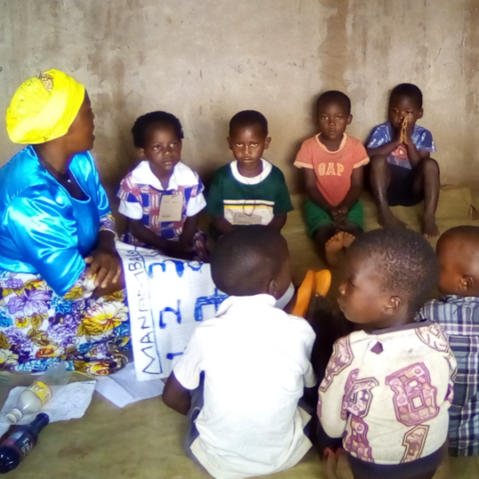 Several African children sitting on the floor gathered around a teacher with learning materials in her lap