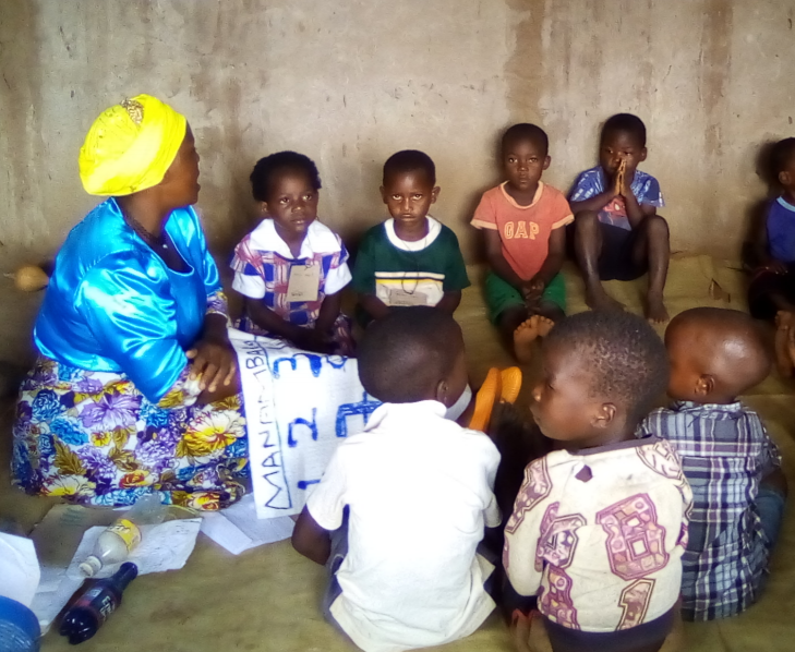 Several African children sitting on the floor gathered around a teacher with learning materials in her lap